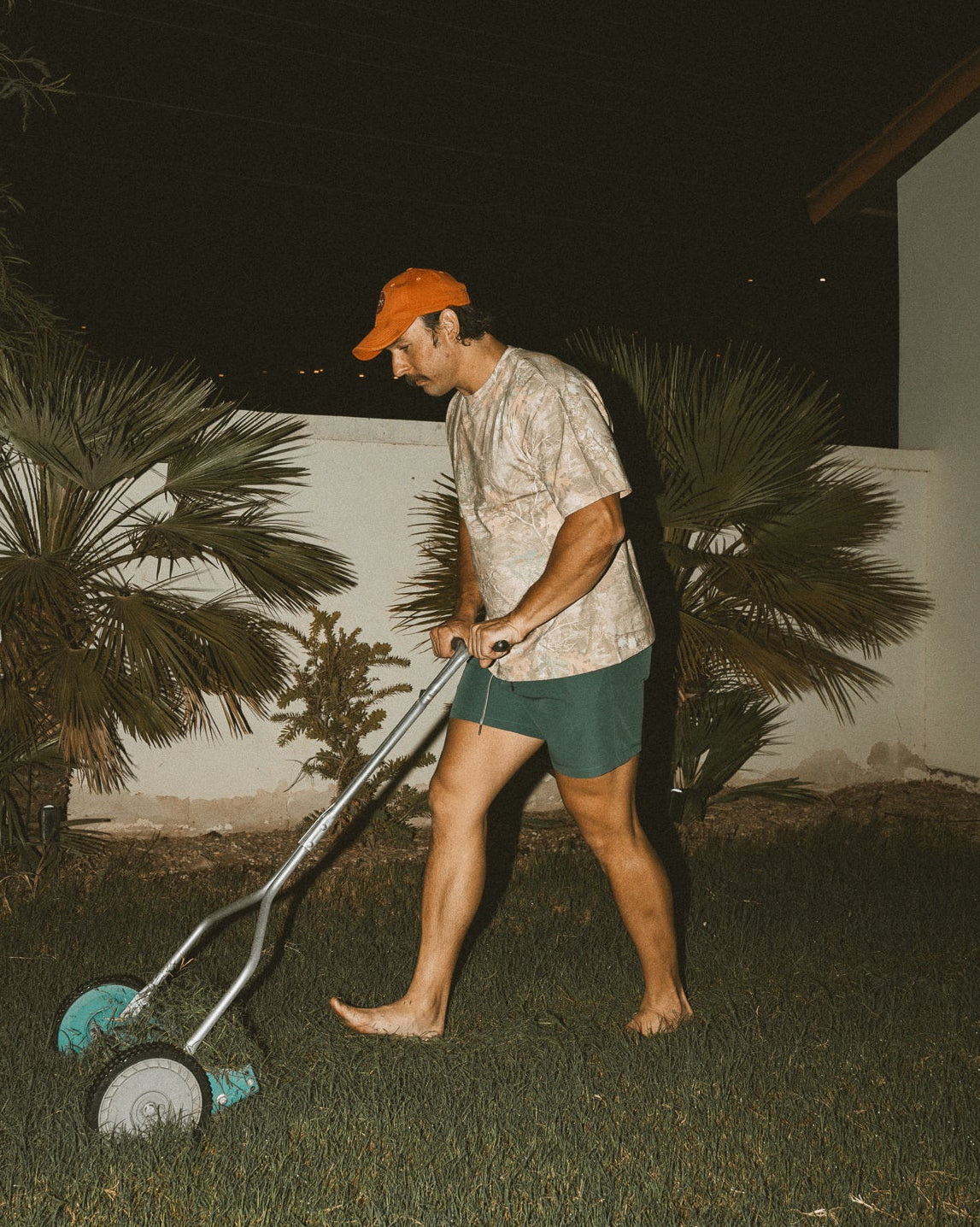 Person using a lawn mower at night in a garden with palm trees.