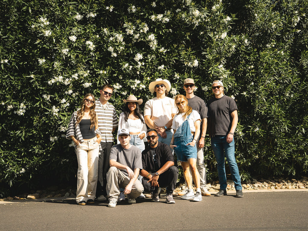 Group of people posing outdoors with a green bush and white flowers in the background